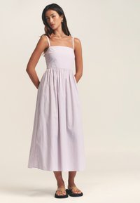 Šviesiai violetinė midi suknelė su vertikaliomis baltomis juostelėmis, aptempta liemenė, kvadratinės iškirptės ir laisvai krentanti suknelės apačia. Dėvima su juodomis šlepetėmis.