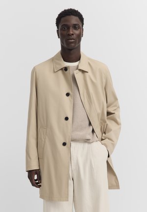 Mann trägt beigen einreihigen Mantel über leichtem Strickpullover und weiten cremefarbenen Hosen, steht mit einer Hand in der Tasche vor einfarbigem Hintergrund.