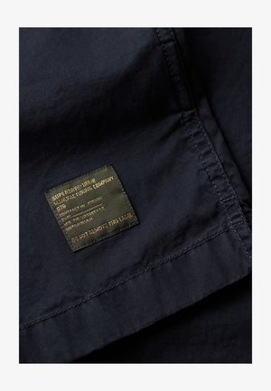 Navy stoffen label met gouden opdruk; bevat merknaam, verzorgingsinstructies en een waarschuwing om niet te verwijderen. Gladde textuur en rechthoekige vorm.