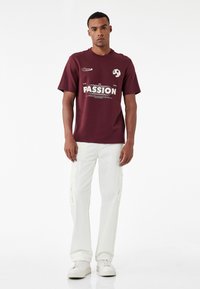 Bordeaux katoenen T-shirt met korte mouwen, featuring witte tekst "LEEF HET LEVEN MET PASSIE" en een klein logo. Gepaarde met witte cargobroeken.