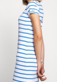 Persona snella in piedi di lato, che indossa un abito bianco con strisce orizzontali blu e il testo "Polo" sulla manica.