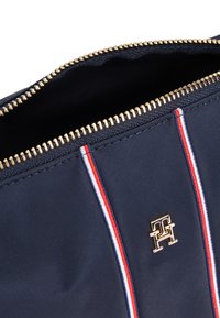 Tamsiai mėlyna nailono krepšys su auksine užtrauktuku ir baltomis, raudonomis bei mėlynomis juostelėmis. Netoli užtrauktuko yra auksinis logotipo akcentas. Audinys turi tekstūrą.