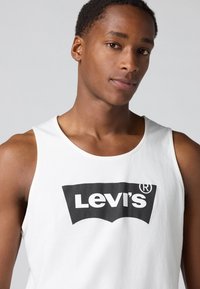 Bijela pamučna majica bez rukava s velikim crnim Levi's logotipom preko prsiju, okruglim dekolteom i širokim izrezima za ruke.