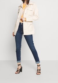 Chaqueta crema de manga larga con dos bolsillos en el pecho, llevada sobre una blusa de estampado, combinada con jeans ajustados azul oscuro y tacones negros con tiras.