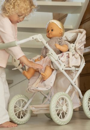 Enfant aux cheveux blonds bouclés jouant avec une poupée assise dans une poussette à motifs à l'intérieur près d'un escalier en bois.