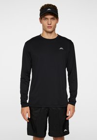 Långärmad svart träningsskjorta med rund halsringning, tillsammans med svarta shorts. Har en liten vit logotyp på skjortan. Slät textur.