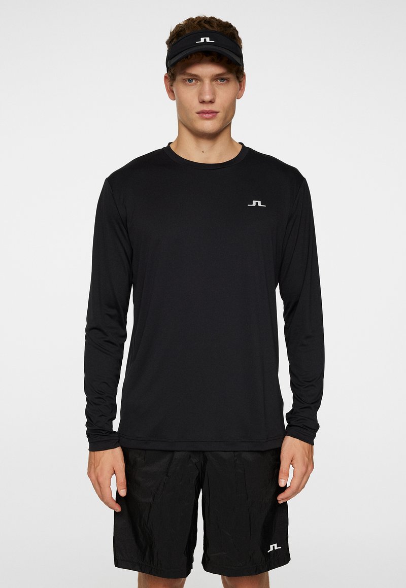 Långärmad svart träningsskjorta med rund halsringning, tillsammans med svarta shorts. Har en liten vit logotyp på skjortan. Slät textur.