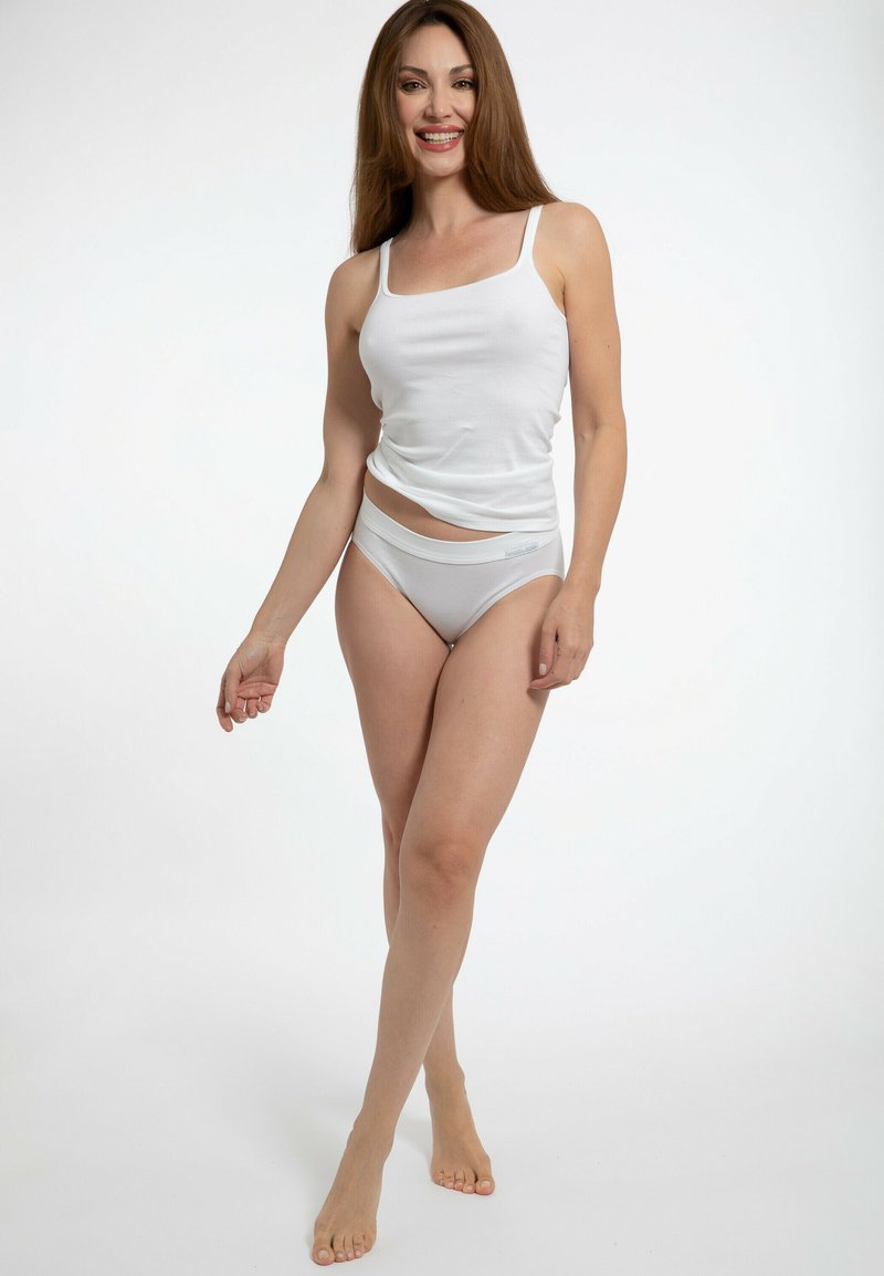 Top in cotone bianco senza maniche con spalline sottili, abbinato a uno slip bikini grigio elasticizzato. Il modello posa a piedi nudi contro uno sfondo semplice.