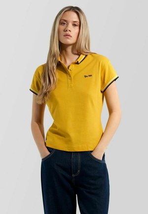 Junge Frau mit langen blonden Haaren, die ein gelbes Poloshirt mit dem Schriftzug "Gabs 1984" und dunkelblaue Jeans trägt, Hände in den Taschen, neutraler Hintergrund.