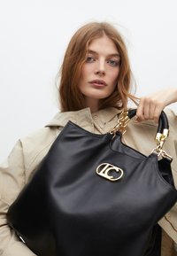 Žena s dlouhými vlnitými vlasy drží černou koženou kabelku se zlatým logem, má na sobě béžovou bundu a stojí na světle šedém pozadí.