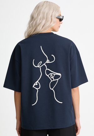 Blonde Person trägt eine Sonnenbrille und ein marineblaues oversized T-Shirt mit einer weißen abstrakten Einlinien-Gesichtzeichnung auf dem Rücken.