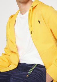 Gul zip-up hoodie med dragsko, med en svart logotyp. Under den finns en vit T-shirt synlig. Marinblå kordflöjlsbyxor med grön randdetalj.