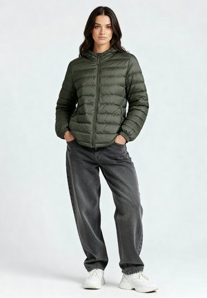 Žena stojí s rukami v kapse, oblečená v olivovo zelenej prešívanej bunde, tmavosivých voľných džínsoch a bielych teniskách na jednofarebnom pozadí.