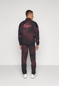 Melna un sarkana sporta tērps ar gradējošu dizainu. Aizmugurē ir izšūts krokodila logo un elastīgas aproces. Baltas sporta kurpes.