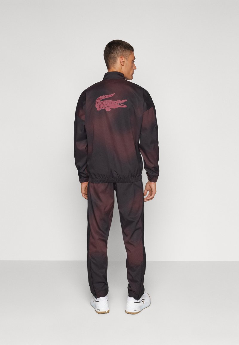 Schwarz-rotes Trainingsanzug mit einem Farbverlauf-Design. Verfügt über ein gesticktes Krokodil-Logo auf dem Rücken und elastischen Bündchen. Weiße Sneakers.