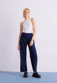 Witte mouwloze top, marineblauwe sportbroek met zijnaad en zwarte klompen. Gladde stof met een relaxte pasvorm en geen zichtbare patronen.