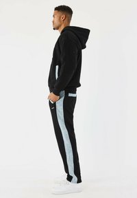 Sudadera negra con bolsillo frontal, combinada con pantalones negros que tienen rayas laterales azul claro y zapatillas blancas. Diseño atlético casual.