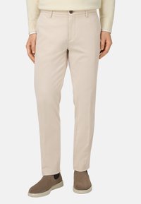Pantalones de algodón beige con un corte recto, que cuentan con un cierre frontal de botón y bolsillos laterales, combinados con zapatos marrones tipo slipping.