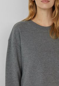 Femme aux longs cheveux blonds portant un pull en tricot gris uni avec un col rond côtelé, sur fond gris clair.