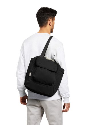 Man wearing white sweatshirt and light pants carries a large black shoulder bag with a front flap pocket.