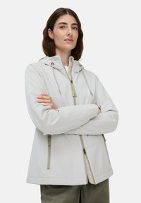 Vrouw met bruin haar, gekleed in een lichtgrijze hoodie met ritszakken, die met gekruiste armen tegen een effen achtergrond staat.