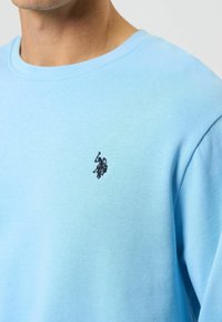 Vaaleansininen puuvillainen collegepaita pyöreällä kaula-aukolla ja pieni musta kirjailtu logo rinnassa. Sileä pinta, rento tyyli.