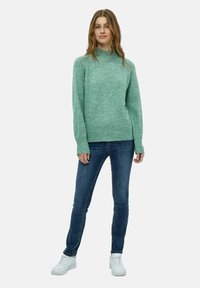 Maglione in lana leggera color teal con collo alto, polsini e orlo a costine, abbinato a jeans blu scuro e sneakers bianche.