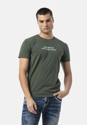 Junger Mann trägt ein olivgrünes T-Shirt mit dem Text "UNLIMITED CIPO AND BAXX" und blaue Jeans, mit einer Hand in der Tasche vor einem schlichten Hintergrund.