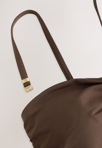 Borsa di stoffa marrone con tracolla sottile e fibbia in metallo dorato su sfondo chiaro, che mostra l’angolo superiore e i dettagli della tracolla.