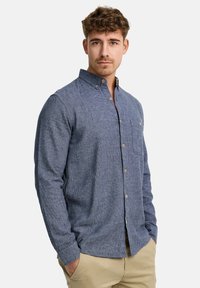 Camisa de manga larga abotonada en tejido texturizado de color azul marino, con un pequeño bolsillo en el pecho y botones de color marrón claro, combinada con pantalones caqui.