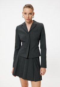 Blazer gris con un solo bolsillo en el pecho, combinado con una falda gris con pliegues. La tela tiene una textura sutil, con mangas largas y un diseño ajustado.