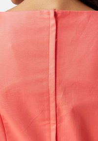 Korálový top s hladkou texturou, se zipem umístěným uprostřed vzadu a rovným výstřihem. Jednoduchý a čistý design.