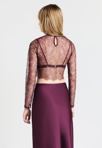 Top in pizzo a maniche lunghe color malva, con un motivo floreale in rilievo, abbinato a una gonna fluida burdeos a tinta unita. Dettaglio di chiusura a forma di chiave sul retro visibile.