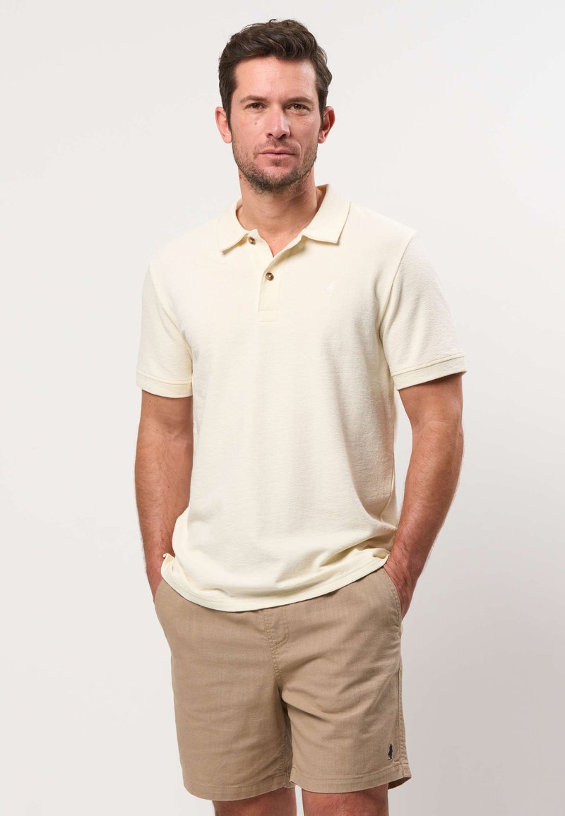 Homme portant un polo crème à manches courtes et un short beige, debout les mains dans les poches devant un fond clair uni.