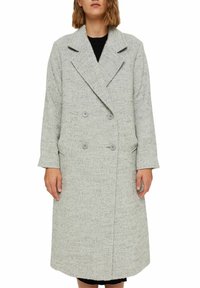 Woman wearing a long, light grey textured double-breasted coat with wide lapels and two side pockets over a black dress.