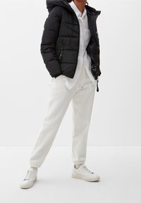 Osoba ubrana w czarną kurtkę puchową, białą koszulę na guziki, teksturowane białe dresy i białe sneakersy na jednolitym tle.