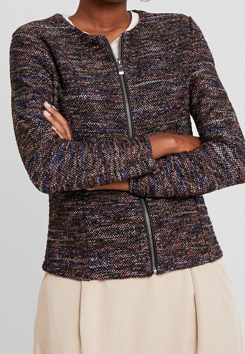 Personne aux bras croisés portant une veste zippée multicolore texturée et une jupe beige clair sur un fond gris uni.