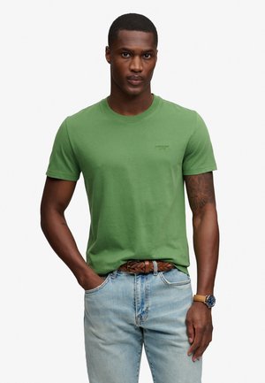 Mann trägt grünes T-Shirt mit Rundhalsausschnitt, hellblaue Jeans mit braunem geflochtenem Gürtel und eine Uhr mit braunem Lederarmband, steht mit einer Hand in der Tasche.