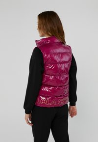 Magenta glanzende puffer-vest met een hoge kraag, voorzien van horizontale stiksels en de opdruk "LOLA CSD" op de rug, gedragen over een zwart shirt.