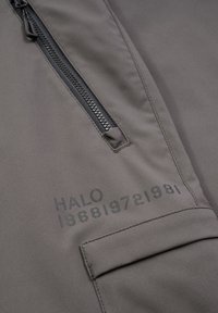 Grå jacka av tyg med en slät textur, med en svart dragkedja, en ficklockficka och tryckta detaljer med texten "HALO 196819721981."