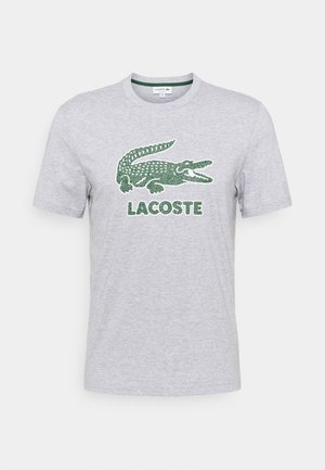 Lysegrå kortærmet t-shirt med stort grønt krokodillelogo og teksten "LACOSTE" centreret på forsiden.