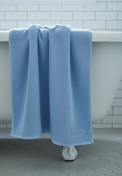 Ανοιχτό μπλε πετσέτα κρεμασμένη στην άκρη μιας λευκής μπανιέρας με πόδια, με λευκές εξαγωνικές πλακάκια στο πάτωμα και λευκά πλακάκια subway στον τοίχο.