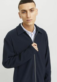 Marineblaue, gerippte Jacke mit Reißverschluss, freiem Kragen, langen Ärmeln und einem silbernen Reißverschluss. Verfügt über einen geschichteten Look mit einem hellen Shirt darunter.