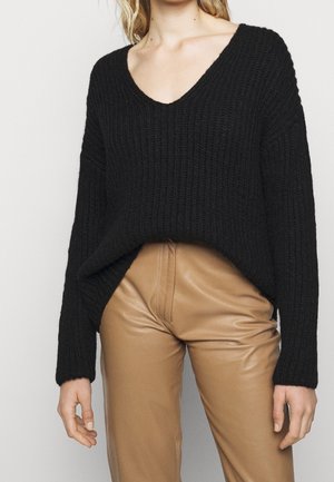 Kvinde iført en sort strikket V-hals sweater og højttaljede tan læderbukser, stående foran en ensartet hvid baggrund.