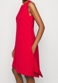 Röd ärmlös klänning med rak skärning, sidofickor och en fåll som kurvar lätt uppåt. Slät tygkänsla.