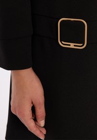 Close-up of a hand next to a black garment with a wide belt featuring a large beige rectangular buckle.