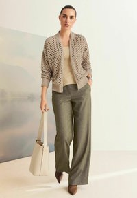 Vrouw in een beige gebreide cardigan, met een tan top, grijze wijde broek en bruine puntige schoenen, die een crèmekleurige handtas vasthoudt in een minimalistische setting.