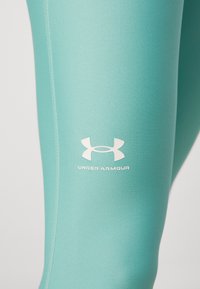 Tirkizne tajice s uskim fitom, napravljene od glatkog materijala. Može se vidjeti bijeli Under Armour logo na butini. Šavovi su vidljivi.