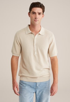 Ung man med kort mörkt hår som bär en beige kortärmad stickad polo och ljusblå jeans, stående mot en enfärgad bakgrund.