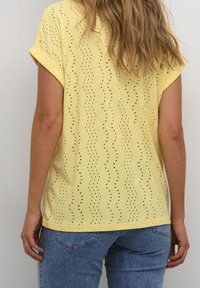 Keltainen lyhythihainen paita, jossa on aaltoilukuosi ja pieniä reikiä. Yhdistetty sinisiin farkkuihin, jotka näyttävät rentoilta.